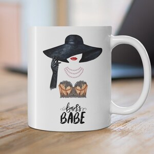 May include: White ceramic mug with a black and white illustration of a woman wearing a black hat and a lace bra. The text "boss babe" is printed in black below the illustration.