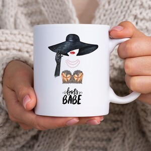 May include: White ceramic mug with a black and white illustration of a woman wearing a black hat and a gold and black lace top. The text "boss babe" is written in black below the illustration.