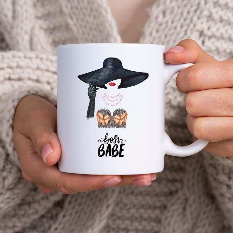 May include: White ceramic mug with a black and white illustration of a woman wearing a black hat and a gold and black lace top. The text "boss babe" is written in black below the illustration.