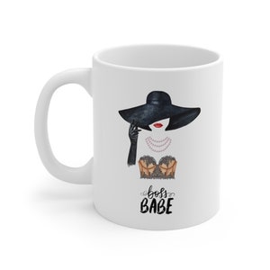 May include: White ceramic mug with a black and white illustration of a woman wearing a black hat, a black lace top, and a pearl necklace. The text "boss babe" is written in black cursive below the illustration.