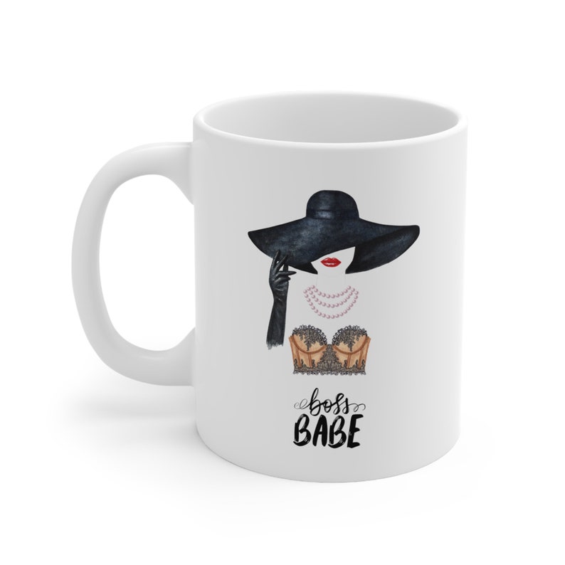 May include: White ceramic mug with a black and white illustration of a woman wearing a black hat, a black lace top, and a pearl necklace. The text "boss babe" is written in black cursive below the illustration.