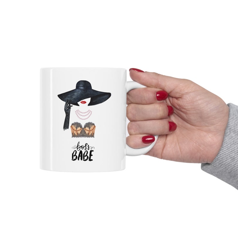 May include: White ceramic mug with a black and white illustration of a woman wearing a black hat, a pearl necklace, and a lacy bra. The text "boss babe" is printed below the illustration.