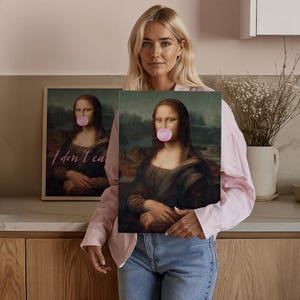 May include: A woman holds two framed prints of the Mona Lisa painting. The Mona Lisa in the print on the right has a pink bubble gum in her mouth. The print on the left has the text "I don't ca" written on it.