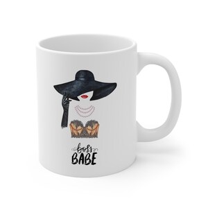 May include: White ceramic mug with a black and white illustration of a woman wearing a black hat, a black lace bra, and a pearl necklace. The text "boss babe" is written in black cursive below the illustration.