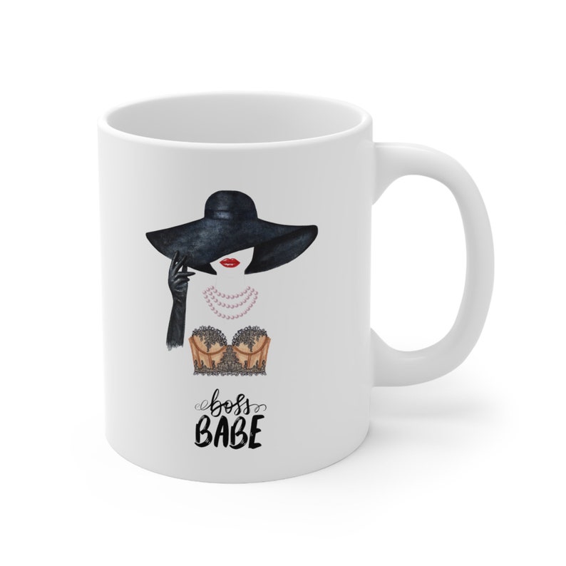 May include: White ceramic mug with a black and white illustration of a woman wearing a black hat, a black lace bra, and a pearl necklace. The text "boss babe" is written in black cursive below the illustration.