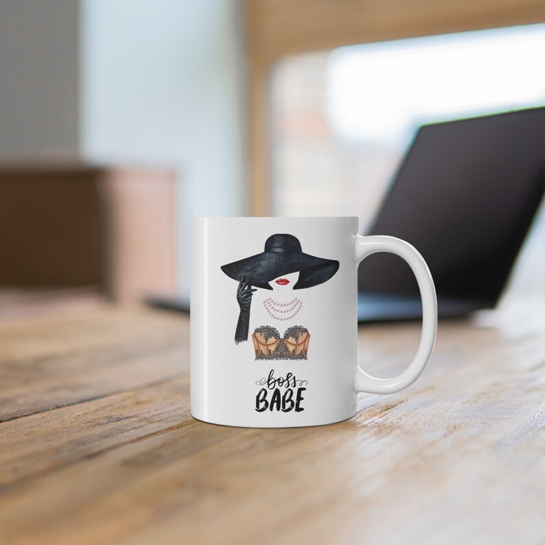 May include: White ceramic mug with a black and white illustration of a woman wearing a black hat and a lace bra. The text "boss babe" is printed in black below the illustration.