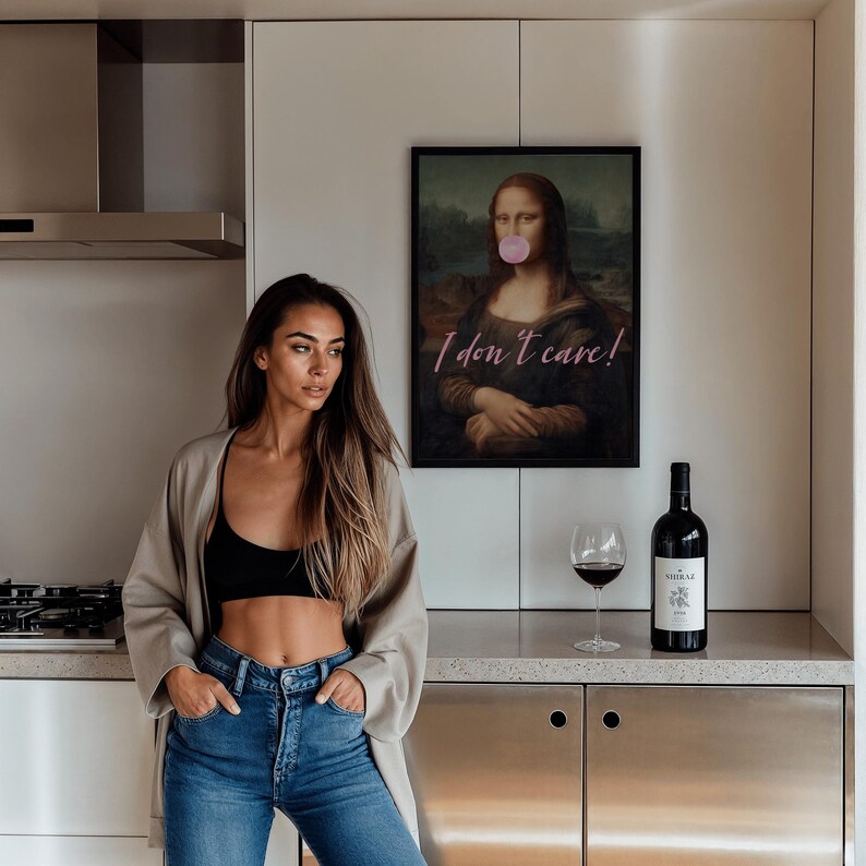 May include: A woman with long brown hair is wearing a beige cardigan, a black bra, and blue jeans. She is standing in a kitchen with a stainless steel counter and cabinets. Behind her is a framed print of the Mona Lisa with a pink bubble gum in her mouth and the text "I don't care!" written in pink.