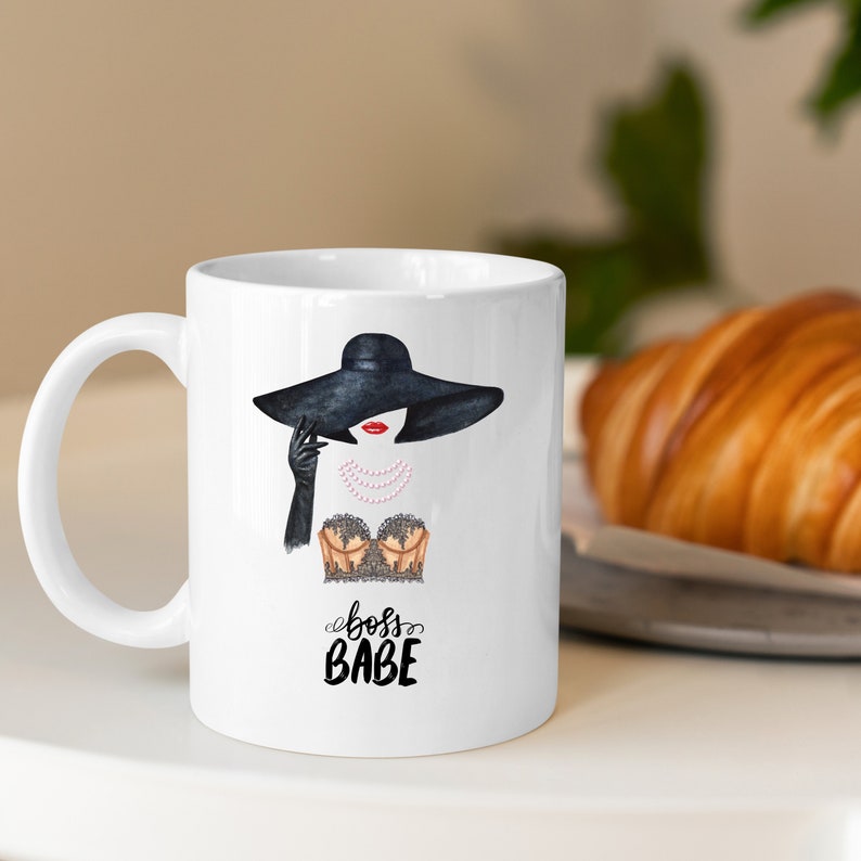 May include: White ceramic mug with a black and white illustration of a woman wearing a black hat and a black and gold top. The text "boss babe" is written in black below the illustration.