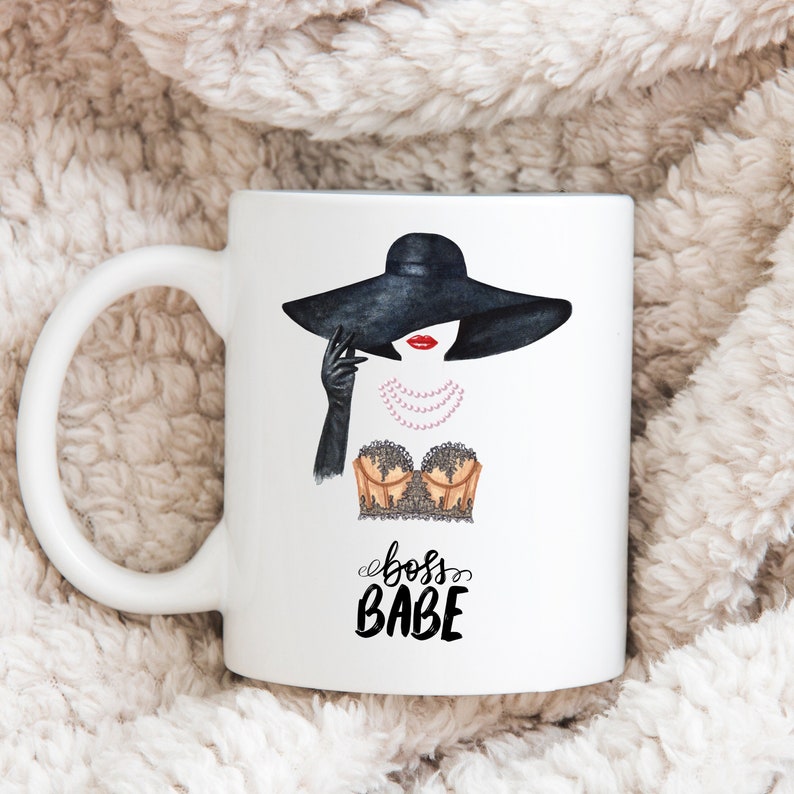 May include: White ceramic mug with a black and white illustration of a woman wearing a black hat and a lace bra. The text "boss babe" is written in black cursive below the illustration.