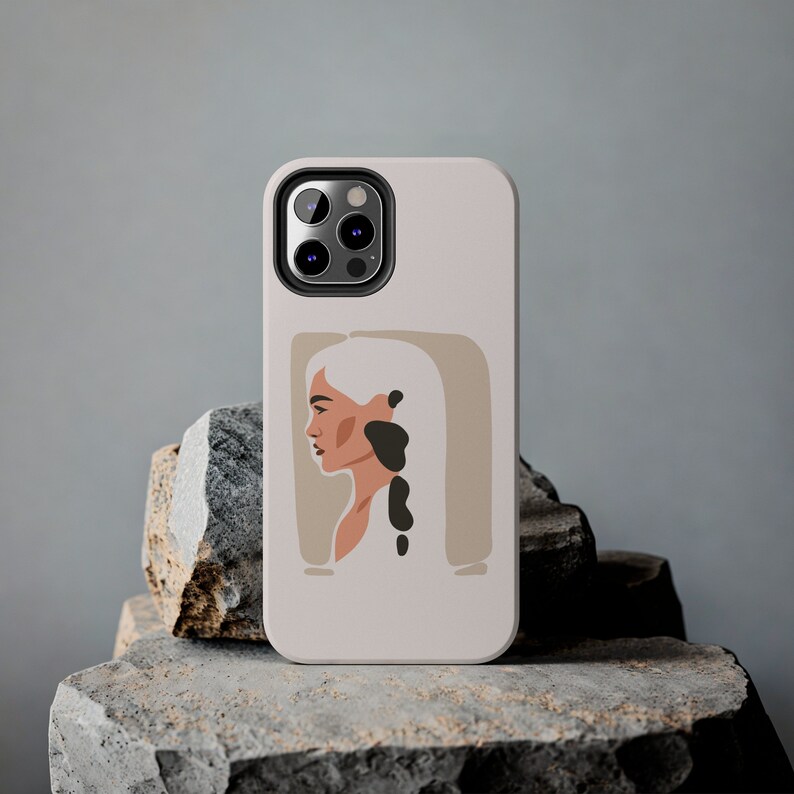 Op de afbeelding: Een witte telefoonhoes met een zwarte camera lens en een minimalistische illustratie van een vrouw met blond haar en een zwart-wit ontwerp. De hoes heeft een beige achtergrond.