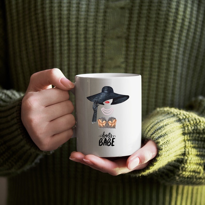 May include: White ceramic mug with a black and white illustration of a woman wearing a black hat and a pearl necklace. The text "boss babe" is printed in black below the illustration.