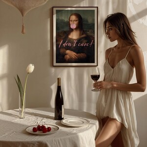 May include: A woman in a white dress holds a glass of red wine. A framed print of the Mona Lisa with the text "I don't care!" is hanging on the wall behind her.