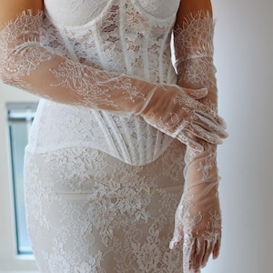 May include: A white lace corset with a strapless neckline and matching lace gloves. The corset has a boned structure and a fitted silhouette.