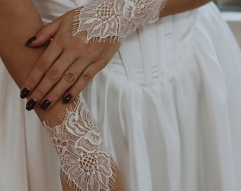 Könnte beinhalten: Ein Paar weiße Spitzen-Handgelenksmanschetten mit einem zarten Blumenmuster. Die Manschetten werden an den Handgelenken einer Person getragen, die ein weißes Kleid trägt.