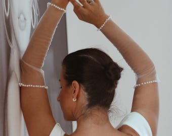 以下が含まれることがあります： 白いオフショルダーのドレスを着た女性が、透き通った真珠で飾られた袖をしています。彼女は白黒の肖像画の前に立っています。