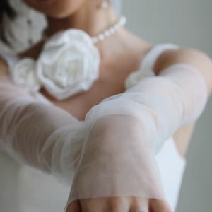 May include: A bride wearing a white wedding dress with a white flower corsage and a sheer white veil. She is wearing a diamond ring on her finger.