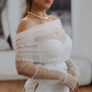 Könnte beinhalten: Weißes Brautensemble mit trägerlosem Oberteil und hochtaillierter Hose. Ein durchsichtiger, schulterfreier Schal und Handschuhe sind mit kleinen Perlen verziert. Eine Perlenkette rundet den Look ab und schafft eine elegante Ästhetik.