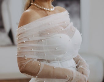 Puede incluir: Conjunto nupcial blanco con corpiño sin tirantes y pantalón de cintura alta. Una envoltura transparente con hombros descubiertos y guantes adornados con pequeñas perlas. Un collar de perlas completa el look, creando una estética elegante.
