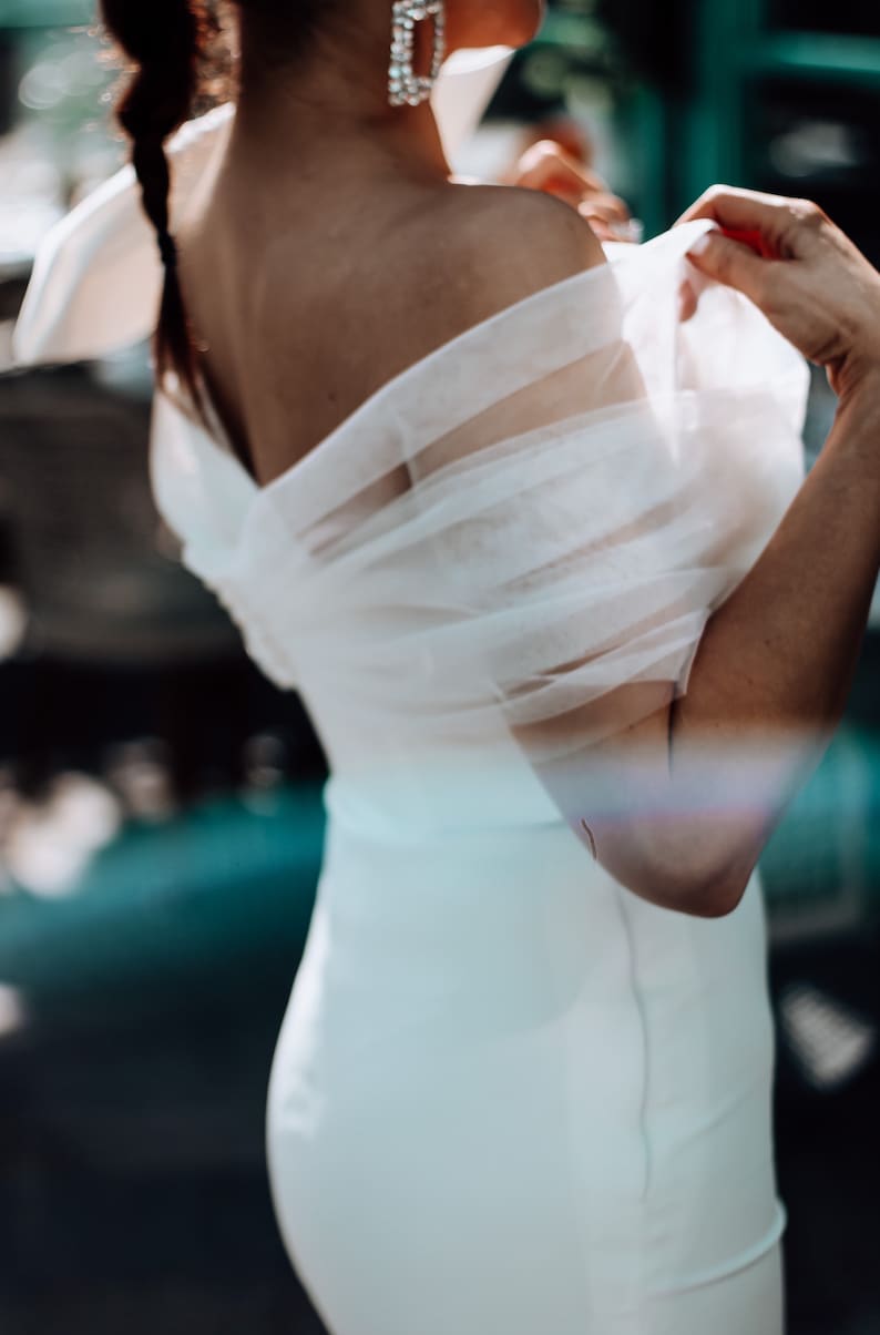 May include: A white off-the-shoulder dress with a sheer overlay. The dress has a fitted bodice and a flowing skirt.
