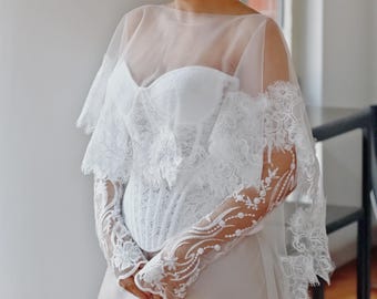 Puede incluir: Un vestido de novia de encaje blanco con una superposición transparente y mangas largas. El vestido tiene un corpiño ajustado y una falda fluida.