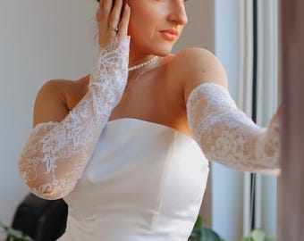 Puede incluir: Una mujer con un vestido de novia blanco sin tirantes y mangas de encaje. Está de pie cerca de una ventana y mirando hacia un lado.