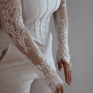 May include: A white corset-style top featuring long, sheer sleeves with intricate white floral lace detailing. The bodice has vertical structural seams and a square neckline, paired with matching white trousers. The lace sleeves extend to the wrists.