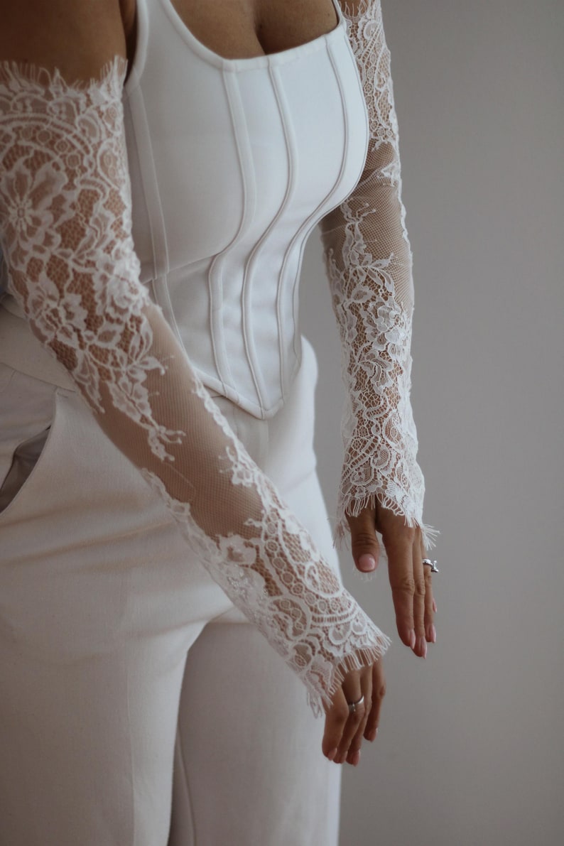 May include: A white corset-style top featuring long, sheer sleeves with intricate white floral lace detailing. The bodice has vertical structural seams and a square neckline, paired with matching white trousers. The lace sleeves extend to the wrists.