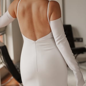 May include: A white wedding dress with a low back and spaghetti straps. The dress has a fitted silhouette that flares out slightly at the bottom. Long, white gloves are worn with the dress. The dress is made of a smooth, likely satin, fabric.