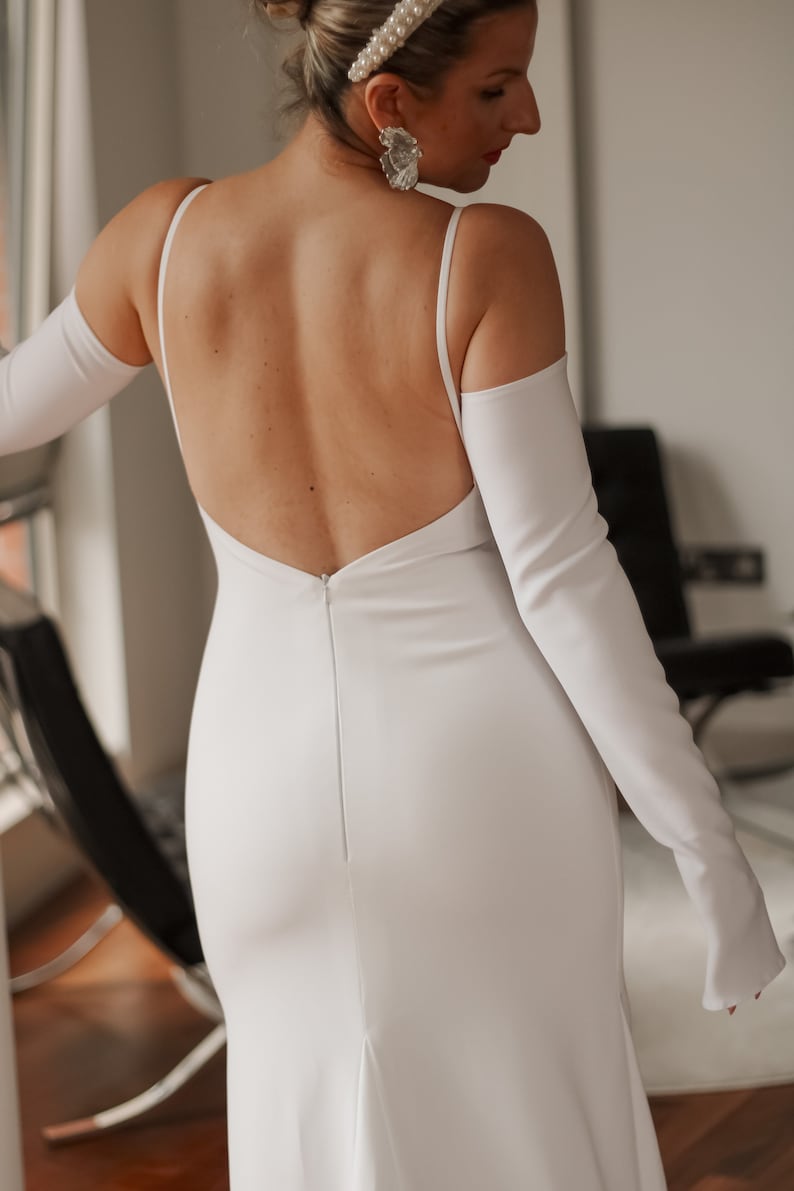 May include: A white wedding dress with a low back and spaghetti straps. The dress has a fitted silhouette that flares out slightly at the bottom. Long, white gloves are worn with the dress. The dress is made of a smooth, likely satin, fabric.