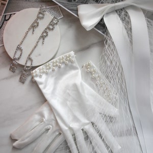 May include: A pair of white satin gloves with pearl trim. The gloves are lying on a white marble surface with a white ribbon and veil.