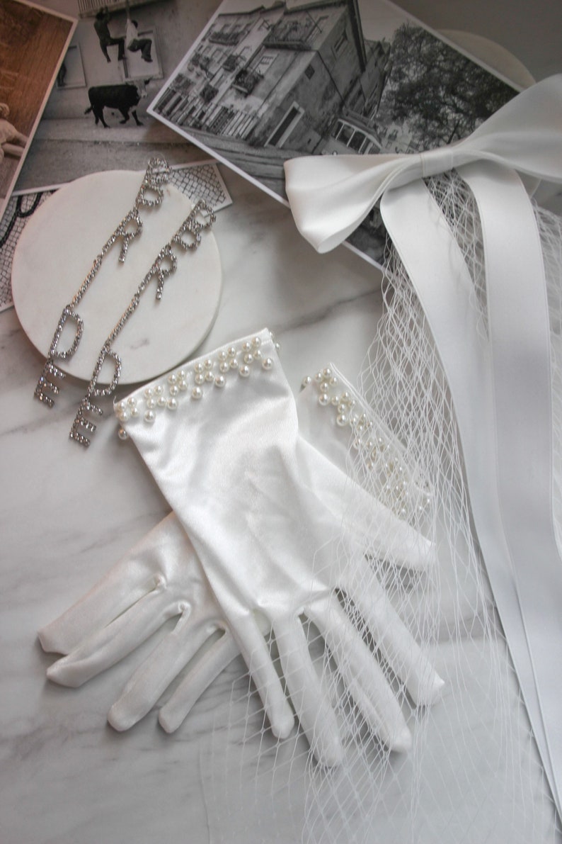 May include: A pair of white satin gloves with pearl trim. The gloves are lying on a white marble surface with a white ribbon and veil.