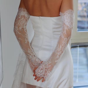 May include: A white strapless wedding dress with a lace overlay and a tulle skirt. The dress has long lace sleeves that are gathered at the wrists.