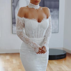 Può includere: Un abito da sposa bianco in pizzo con spalle scoperte, maniche lunghe e un corpetto aderente.