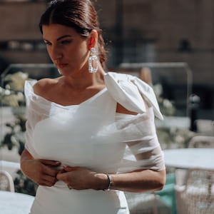 May include: A woman wearing a white off-the-shoulder dress with a large bow on one shoulder. She is standing in front of a cafe with tables and chairs.