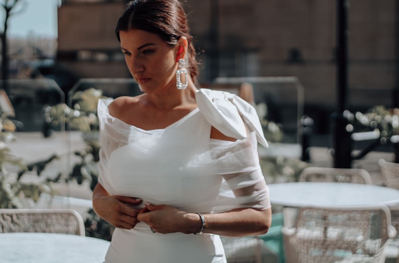 May include: A woman wearing a white off-the-shoulder dress with a large bow on one shoulder. She is standing in front of a cafe with tables and chairs.
