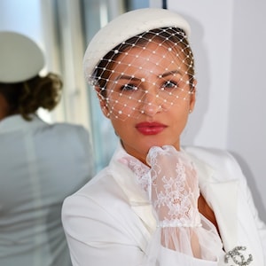 Puede incluir: Un sombrero tipo pastillero blanco con un delicado velo de jaula adornado con pequeñas perlas blancas. Se combina con guantes de encaje blanco con puños de volantes y un broche plateado con un logotipo entrelazado en la solapa de un blazer.