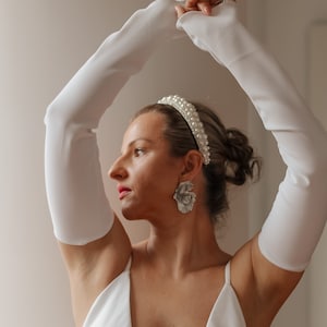 May include: A woman wearing a white dress, long white gloves, a pearl headband, and large silver flower earrings. The woman is posing with her arms raised. The background is a plain, light-colored wall.