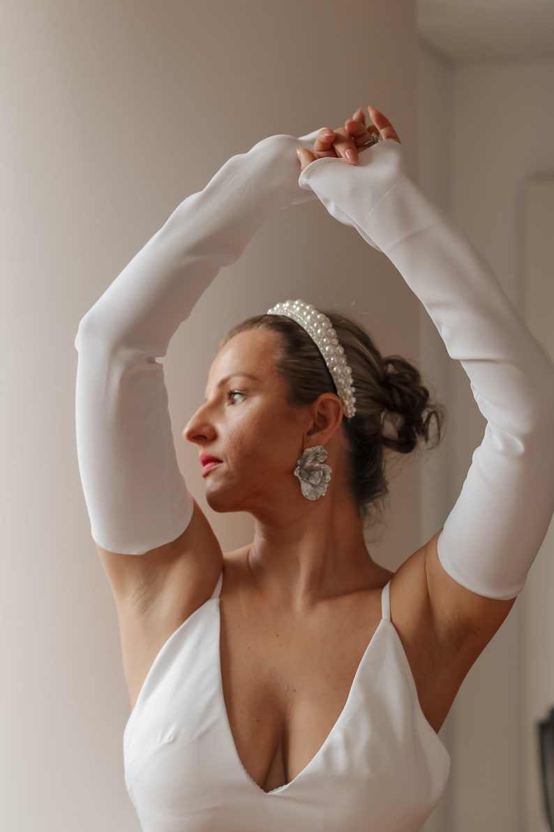 May include: A woman wearing a white dress, long white gloves, a pearl headband, and large silver flower earrings. The woman is posing with her arms raised. The background is a plain, light-colored wall.