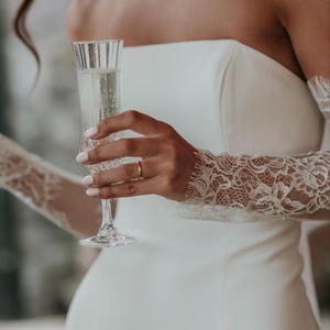 May include: A strapless white gown featuring long, sheer lace sleeves with intricate floral patterns and scalloped edges. The garment is paired with a tall, fluted glass containing a sparkling beverage, held by a hand with a simple gold band.
