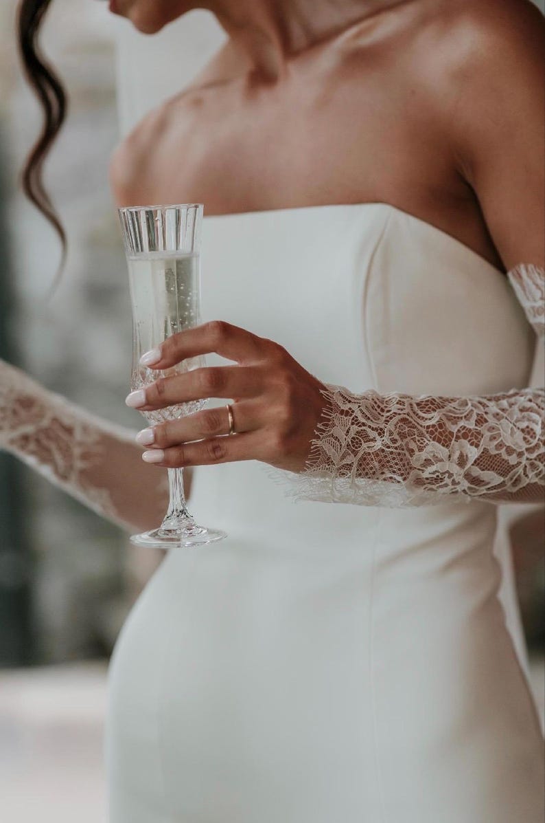 May include: A strapless white gown featuring long, sheer lace sleeves with intricate floral patterns and scalloped edges. The garment is paired with a tall, fluted glass containing a sparkling beverage, held by a hand with a simple gold band.