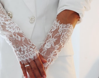 Op de afbeelding: Close-up van witte kanten handschoenen met ingewikkelde bloempatronen. De handschoenen worden gedragen met een witte blazer, versierd met parelachtige knopen. De kanten mouwen voegen een vleugje elegantie toe.
