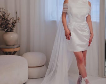 Puede incluir: Un vestido blanco de satén corto con una superposición de tul transparente y una silueta ajustada. El vestido tiene un escote redondo y mangas cortas. La modelo lleva calcetines blancos y tacones.