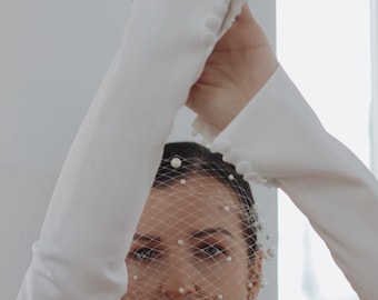 以下が含まれることがあります： 顔に真珠のアクセントが付いた白いベールをつけた女性。白い手袋と真珠のネックレスをしています。