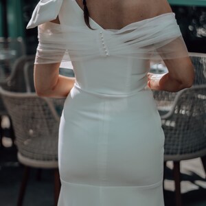 May include: A white off-the-shoulder wedding dress with a tulle overlay and a fitted skirt.