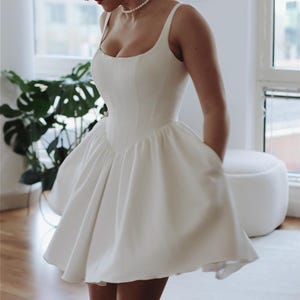 May include: A white, sleeveless, short wedding dress with a fitted bodice and a full skirt. The dress has a square neckline and a simple design.