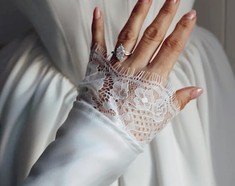 Könnte beinhalten: Ein weißes Satin-Brautkleid mit einer Spitzenborte am Ärmel. Eine Hand trägt einen silbernen Ring mit einem großen, klaren, markisenförmigen Diamanten.