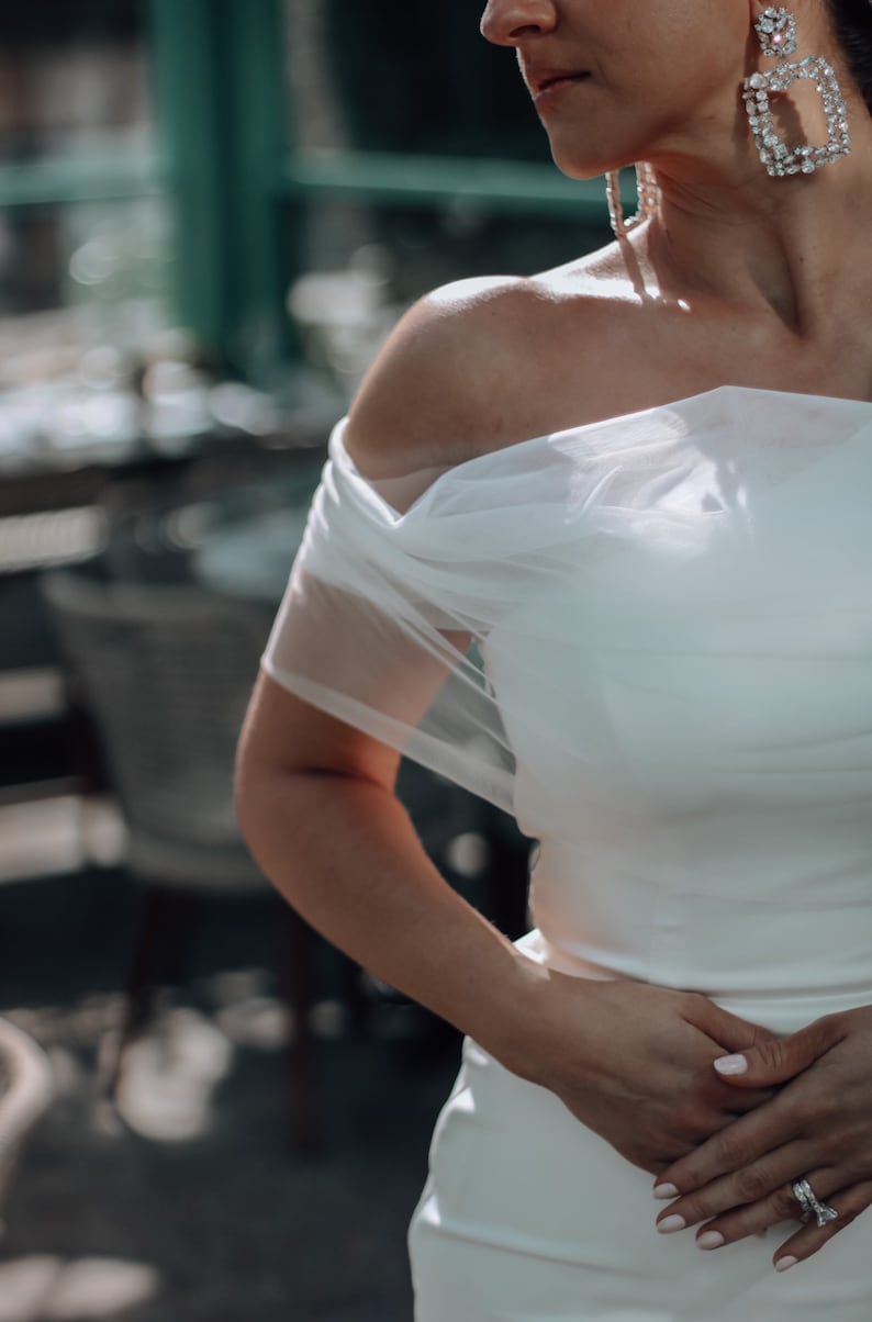 May include: A white off-the-shoulder wedding dress with a sheer overlay. The dress has a fitted bodice and a flowing skirt. The bride is wearing a diamond ring on her finger.