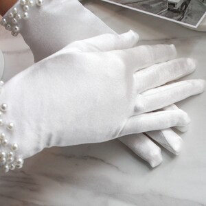 May include: A pair of white satin gloves with pearl embellishments on the wrist. The gloves are lying on a white marble surface.