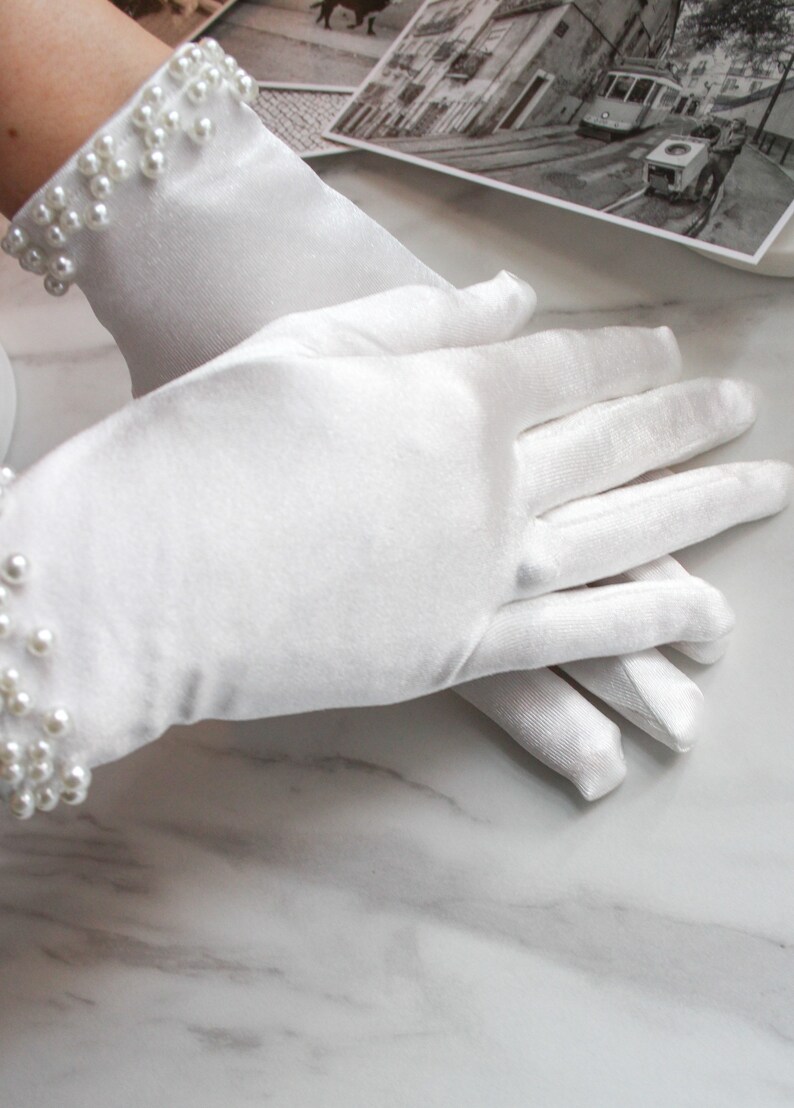 May include: A pair of white satin gloves with pearl embellishments on the wrist. The gloves are lying on a white marble surface.