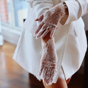 May include: Delicate white lace gloves with detailed floral patterns. The gloves are worn with a white blazer. The intricate lace design covers the entire glove, creating an elegant look. The image is well-lit, highlighting the texture and detail.